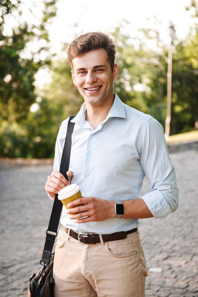 Smiling young man in shirt carrying bag walking at the city park with cup of coffee - Photo, image