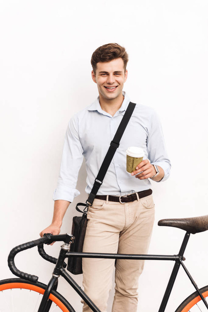 Satisfied young stylish man dressed in shirt carrying bag standing with a bicycle holding coffee cup over white street wall - Fotoğraf, Görsel