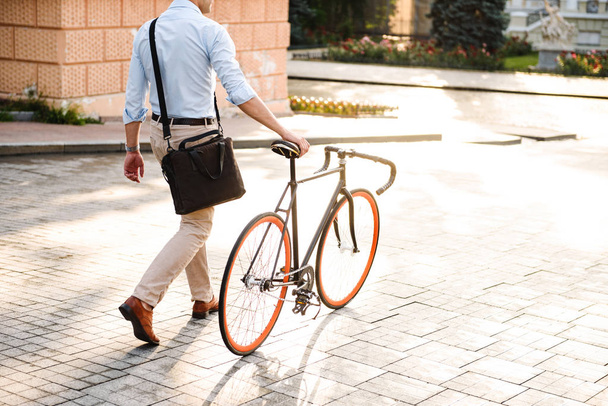Cropped image back view of young stylish man dressed in shirt walking with a bicycle on a city street - Foto, imagen