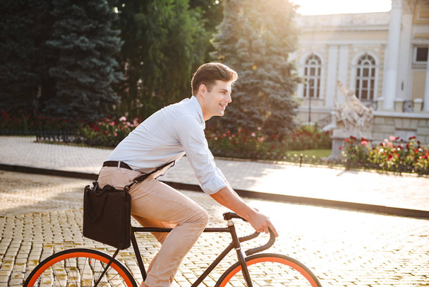 Attractive young stylish man dressed in shirt riding on a bicycle on a city street - Фото, изображение