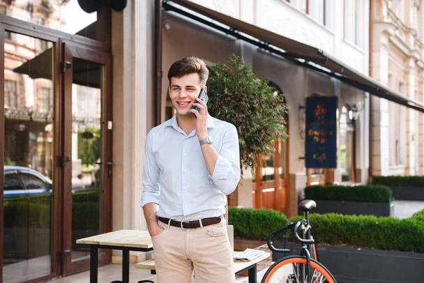 Smiling young stylish man in shirt talking on mobile phone while standing at a cafe outdoors with bicycle - Foto, immagini