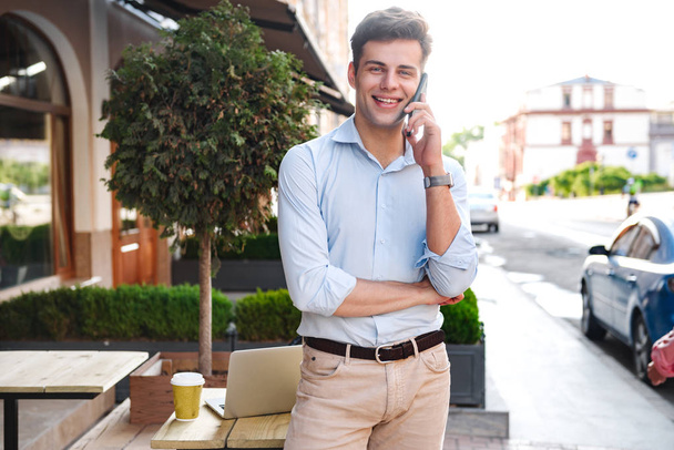 Smiling young stylish man in shirt talking on mobile phone while standing at a cafe outdoors - Foto, Bild