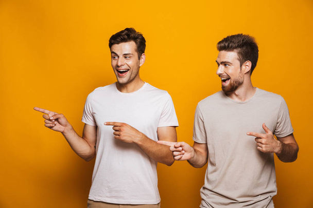 Portrait of a two happy young men best friends pointing fingers away at copy space isolated over yellow background - Photo, Image