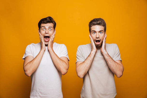 Portrait of a two shocked young men best friends looking at camera with mouth open isolated over yellow background - 写真・画像