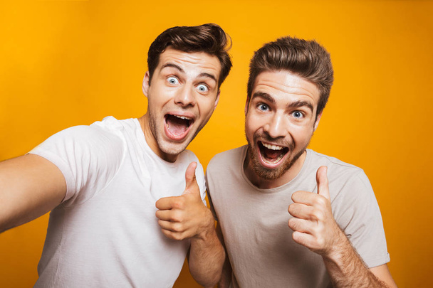 Portrait of a two happy young men best friends showing thumbs up taking a selfie isolated over yellow background - Fotografie, Obrázek