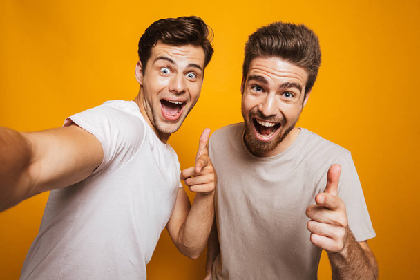 Portrait of a two happy young men best friends pointing fingers at camera taking a selfie isolated over yellow background - Valokuva, kuva