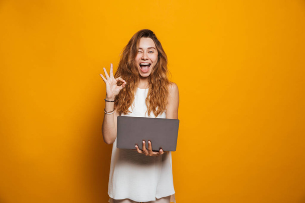 Portrait of a cheerful young girl holding laptop computer and showing ok isolated over yellow background - Zdjęcie, obraz