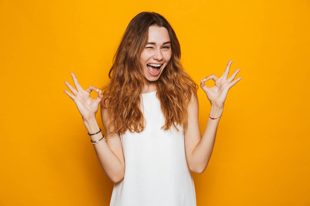 Portrait of a happy young girl looking at camera, showing ok gesture isolated over yellow background - Foto, Imagem