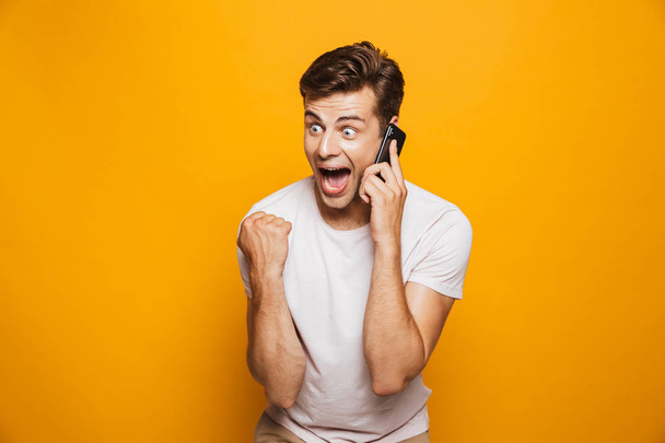 Portrait of a happy young man talking on mobile phone isolated over yellow background, celebrating - Foto, immagini