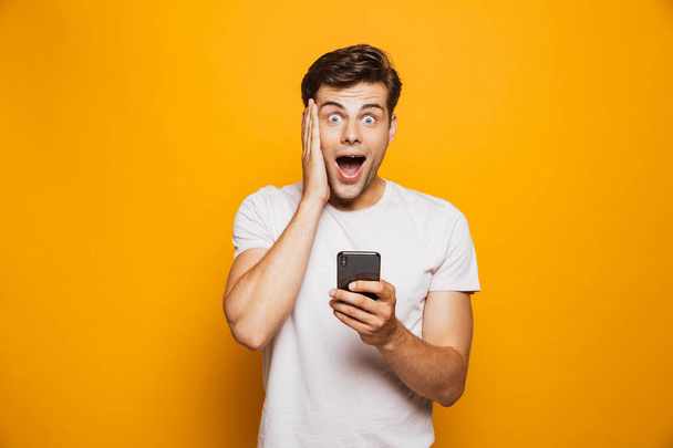Portrait of an astonished young man holding mobile phone isolated over yellow background, celebrating - Foto, Imagen