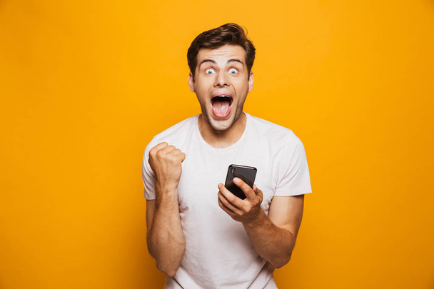 Portrait of a happy young man holding mobile phone isolated over yellow background, celebrating - Fotó, kép