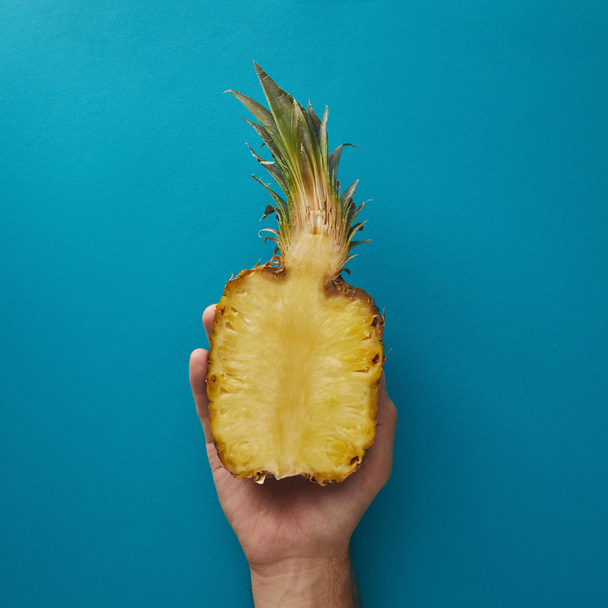 cropped image of man holding half of ripe pineapple above blue surface - Foto, Imagen