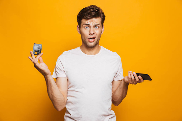 Portrait of a puzzled young man holding mobile phone and showing credit card isolated over yellow background - Foto, afbeelding