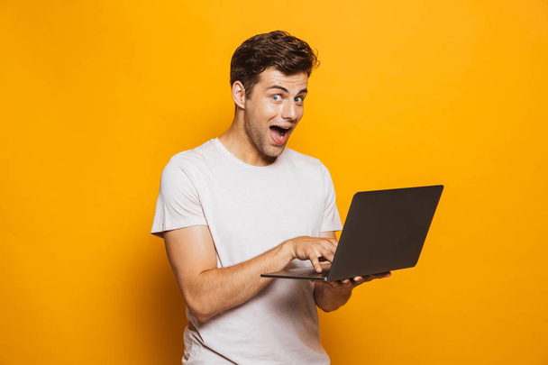 Portrait of an excited young man using laptop computer isolated over yellow background - Fotografie, Obrázek