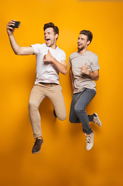 Full length portrait of a two happy young men best friends taking a selfie while jumping isolated over yellow background - Fotó, kép