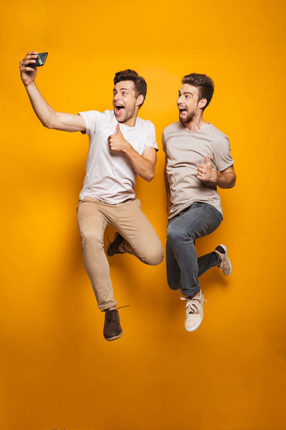 Full length portrait of a two excited young men best friends taking a selfie while jumping isolated over yellow background - Foto, immagini