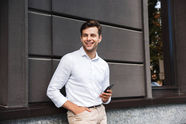Portrait of a smiling young formal dressed man leaning on a wall outdoors holding mobile phone - Fotografie, Obrázek