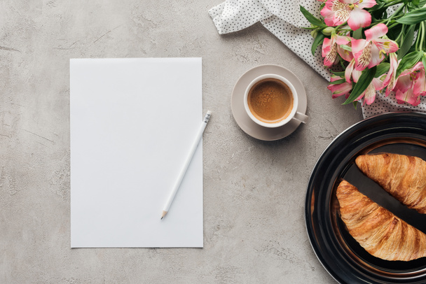 Draufsicht auf Tasse Kaffee mit leerem Papier, Croissants auf Teller und Alstroemeria-Bouquet auf Betonoberfläche - Foto, Bild