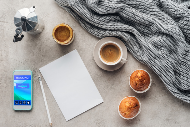 bovenaanzicht van een kopje koffie met muffins, blanco papier en smartphone met boeken app op scherm op betonnen oppervlak met gebreide wol gordijnen - Foto, afbeelding