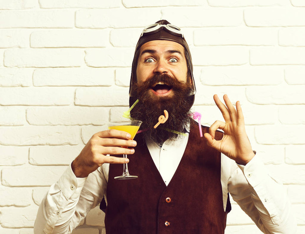 handsome bearded aviator man with long beard and mustache on happy face holding glass of alcoholic cocktail in vintage suede leather waistcoat with hat and glasses on white brick wall background - Photo, image
