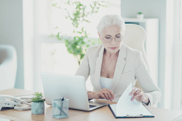 Grey-haired beautiful stylish clever old serious lady boss in glasses sitting in front of netbook, working with paper documents, work place work station - Fotoğraf, Görsel