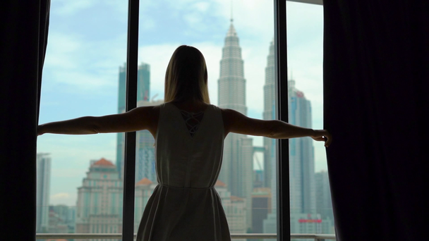 Superslowmotion shot van een silhouet van een succesvolle rijke vrouw openen de gordijnen van een venster met uitzicht op het centrum van de stad met wolkenkrabbers. - Video
