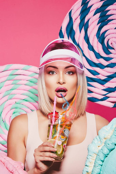 blonde woman wearing top and pink cap posing on pink background with huge sweet lollypops,  drinking lemonade - 写真・画像