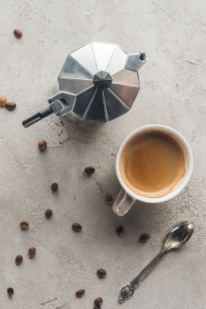 top view of cup of delicious coffee and moka pot on concrete surface with spilled coffee beans - Photo, Image