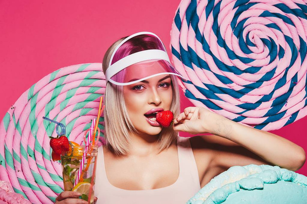 レモネードを押しながらイチゴを食べて、ピンクの背景の先端とピンクのキャップの立っている身に着けているブロンドの髪を持つ美しい少女 - 写真・画像