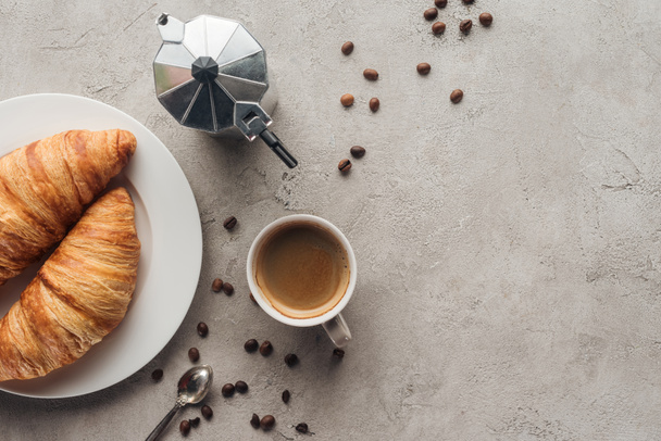 top view of cup of coffee with croissants and moka pot on concrete surface with spilled coffee beans - Photo, Image