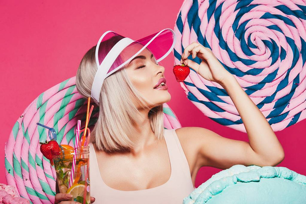 блондинка в верхней и розовой кепке стоит на розовом фоне, ест клубнику
.  - Фото, изображение