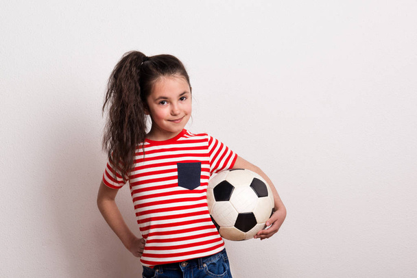 A small girl with a soccer ball and striped T-shirt standing in a studio. - Fotó, kép