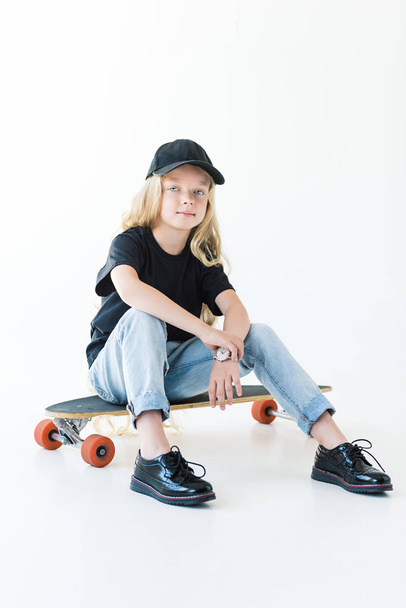 vista completa de niño en gorra negra y camiseta sentado en longboard y mirando a la cámara aislada en blanco
 - Foto, imagen