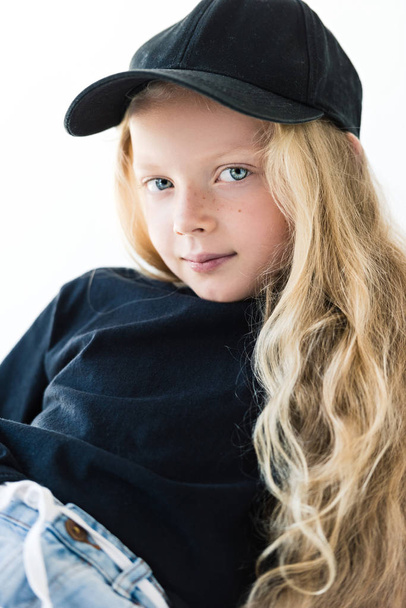 portrait of beautiful child with long curly hair wearing black t-shirt and cap, looking at camera isolated on white - 写真・画像