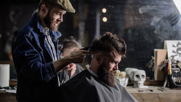 Barber with clipper trimming hair on nape of client. Barber with hair clipper works on haircut of bearded guy barbershop background. Hipster hairstyle concept. Hipster client getting haircut. - Foto, Imagem