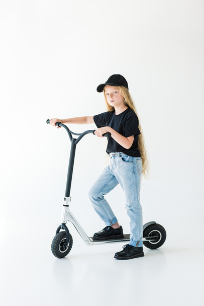 cute stylish child in black t-shirt and cap riding scooter and looking at camera isolated on white   - Fotó, kép