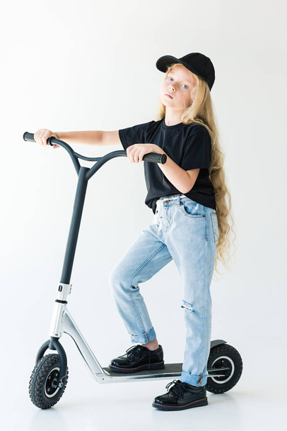vue pleine longueur de l'enfant en t-shirt noir et chapeau scooter équitation et en regardant la caméra isolée sur blanc
  - Photo, image