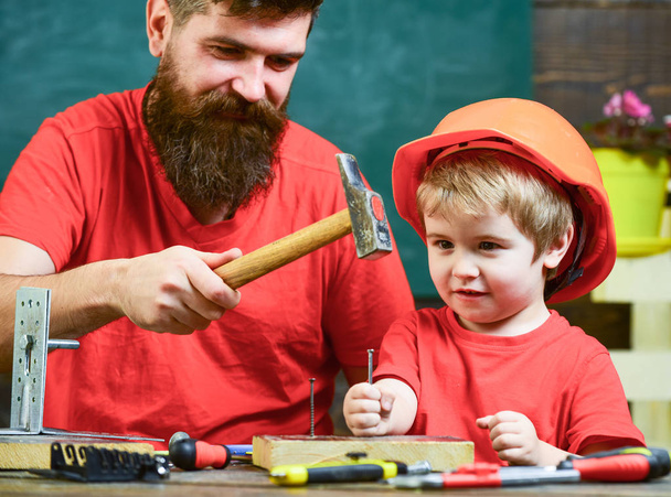 Boy, child busy in protective helmet learning to use hammer with dad. Father with beard teaching little son to use tools, hammering, chalkboard on background. Handyman concept - Foto, Bild