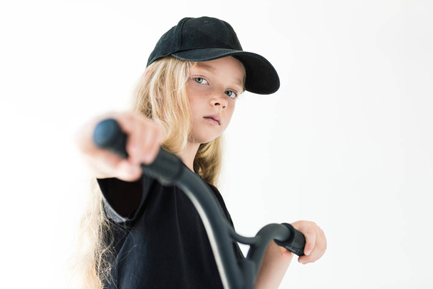 beautiful kid in black t-shirt and cap riding scooter and looking at camera isolated on white  - 写真・画像