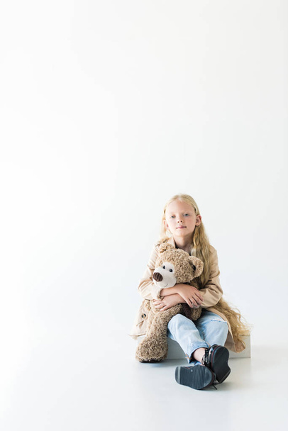 full length view of beautiful little child holding teddy bear and smiling at camera while sitting isolated on white - Photo, Image
