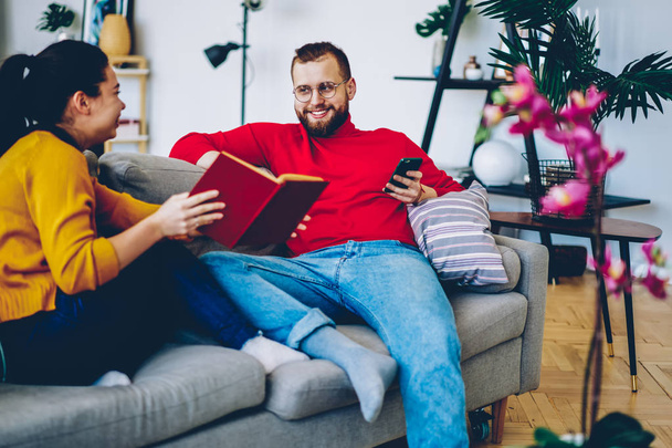Smiling romantic couple discussing new book spending free time together in cozy home interior, mariggecommunicating on leisure, hipster guy holding smartphone while talking to positive girlfriend - Foto, Bild