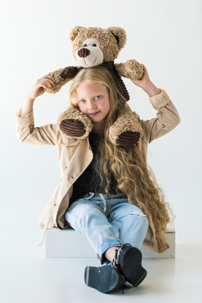full length view of beautiful stylish kid holding teddy bear and smiling at camera isolated on white  - Foto, immagini