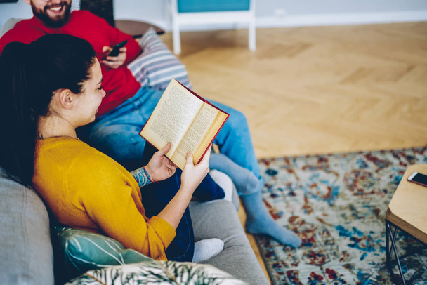 Cropped image of female sitting with book next to boyfriend on sofa at home, couple in love spending time together in apartments , woman holding open bestseller  - Foto, Imagem