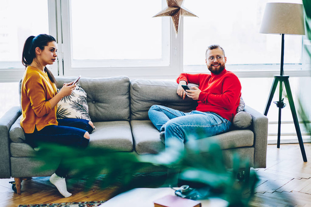 Positive millennial hipster couple sitting on sofa at home interior satisfied with good wireless connection,young family resting on weekends at apartment using smartphone for networking ignore talk - Photo, Image