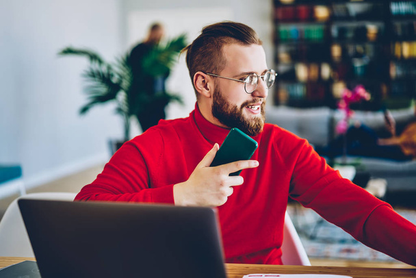 Positive male freelancer satisfied with remote job and wireless connection for using gadgets, handsome millennial male holding smartphone for making telephone call doing distance job and bankin - Photo, image