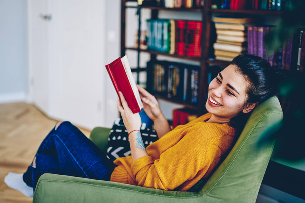 Cheerful brunette woman laughing at funny story from book satisfied with free time at home interior with literature collection, positive young hipster girl enjoying relaxation with interesting novel - Photo, image