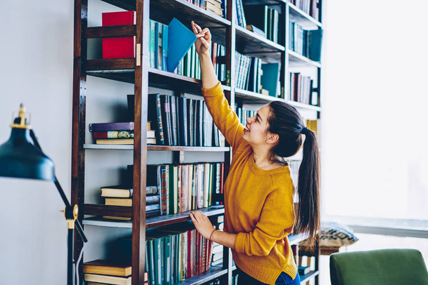 Smiling brunette hipster girl choosing literature from shelves with home collection in living room, positive young woman checking interesting novel from bookcase for spending leisure time on hobb - Foto, Imagem