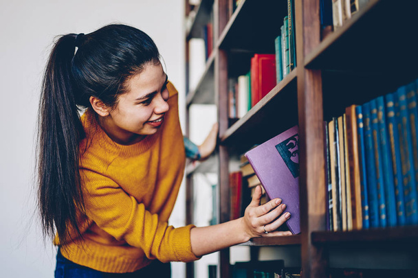 Young caucasian young woman with brunette hair  taking boor for reading on free time standing near bookcase, positive teen hipster girl selecting literature taking novels from shelves in library - Photo, Image