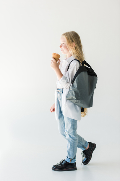 vista lateral de lindo niño sonriente con mochila comiendo helado y mirando hacia otro lado en blanco
 - Foto, imagen