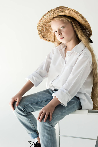 krásné malé dítě v proutěné klobouk sedí a při pohledu na fotoaparát na bílém  - Fotografie, Obrázek